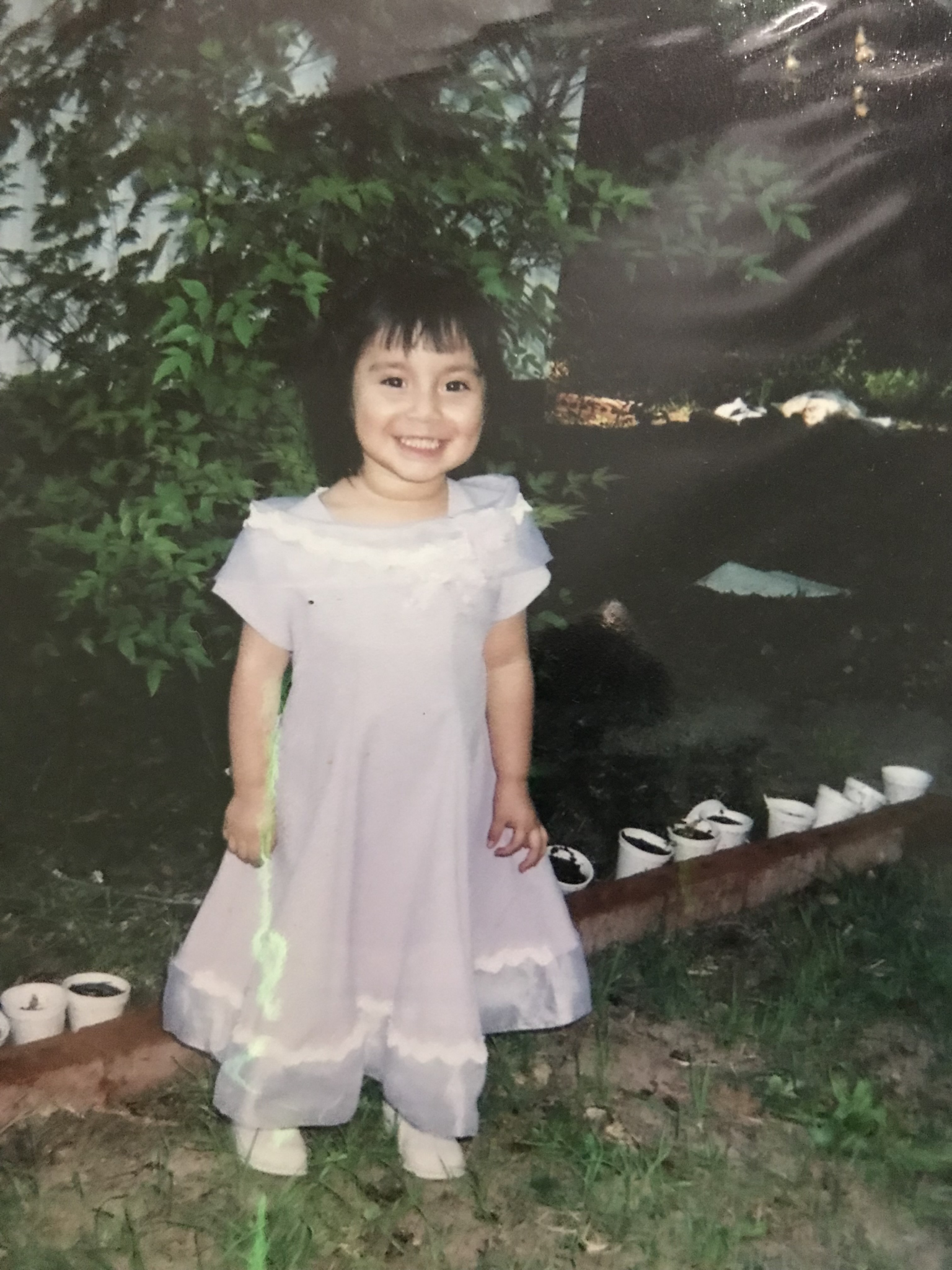 Pre-school age Stephanie stands in front of a garden with a short dark bob and a beautiful white frilly dress.