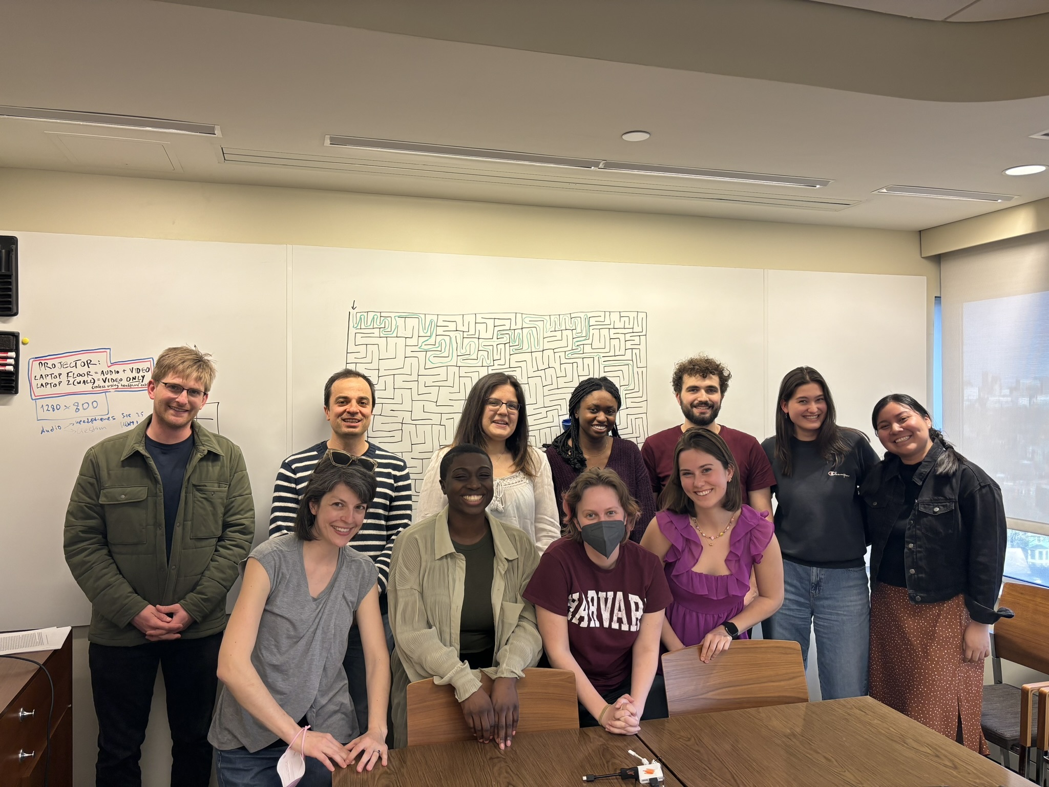Staff, grad student, postdocs, and undergraduate members of the Bergelson lab stand smiling in a classroom with a maze. The image changes to a silly one when the mouse rolls over it.
